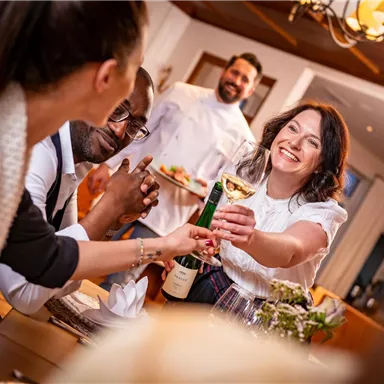 Eine fröhliche Gruppe von Personen stößt mit Gläsern an. Im Hintergrund lächelt ein Koch und serviert Speisen.
