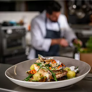 Ein köstlicher Fischgericht mit frischen Kräutern und Zitrone in einem schönen Teller. Im Hintergrund arbeitet ein Koch in einer modernen Küche.