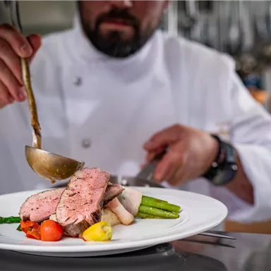 Ein Koch präsentiert ein kunstvoll angerichtetes Gericht mit gebratenem Fleisch und frischem Gemüse. Der Fokus liegt auf der Zubereitung und der Ästhetik des Essens.