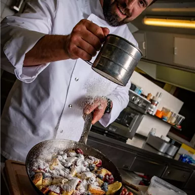Ein Koch streut Puderzucker über eine fruchtige Dessertplatte. Im Hintergrund sind Küchenutensilien sichtbar.