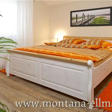 Ein gemütliches Schlafzimmer mit einem großen Bett und orangefarbenen Kissen. Im Hintergrund sind ein Fenster und ein Bild an der Wand zu sehen.