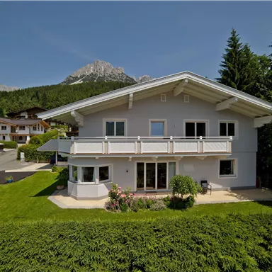 Ein modernes Haus mit weißer Fassade und Balkon, umgeben von einer grünen Wiese. Im Hintergrund sind Berge und Wälder sichtbar.