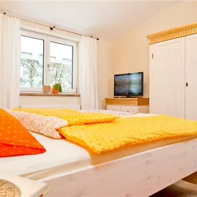 Ein helles Schlafzimmer mit einem großen Bett und bunten Kissen. Im Hintergrund steht ein Fernseher und ein Schrank.