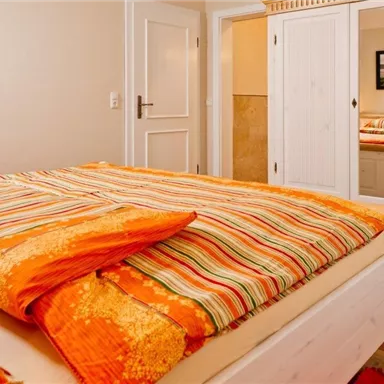Ein gemütliches Schlafzimmer mit einem großen Bett und bunten Bettdecken. Im Hintergrund ist ein Spiegel und eine Tür zu sehen.