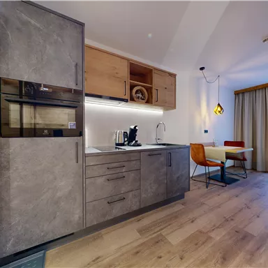 A modern kitchen unit with gray cabinets and wooden details. The room is bright and minimally furnished, with a small dining table and a chair.