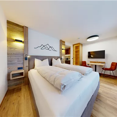 A modern hotel room with a large bed and wood paneling. In the background, a television and a desk can be seen.