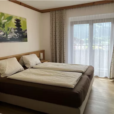 Ein helles Zimmer mit zwei Einzelbetten und Fenster, die viel Tageslicht hereinlassen. An der Wand hängt ein ruhiges Bild mit Pflanzen und Steinen.