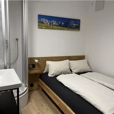 Ein kleines, modernes Zimmer mit einem Doppelbett und beigen Kissen. Auf der linken Seite befindet sich eine Dusche und ein Waschbecken.