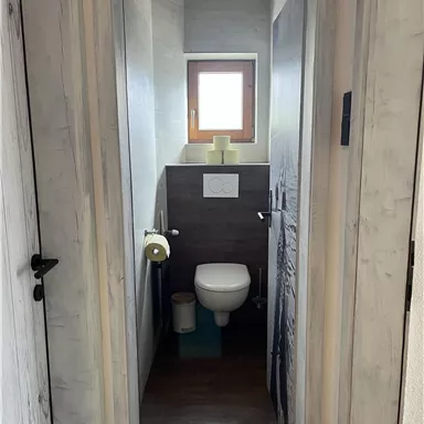 Ein kleines Badezimmer mit einer weißen Toilette und einem Fenster. Die Wände sind in neutralen Tönen gehalten und der Boden ist dunkel.