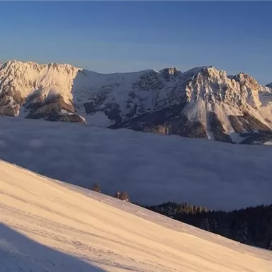 An impressive mountain landscape with snow-covered peaks and a sea of clouds in the valley. The sky is clear and the sun shines on the snow-covered slopes.