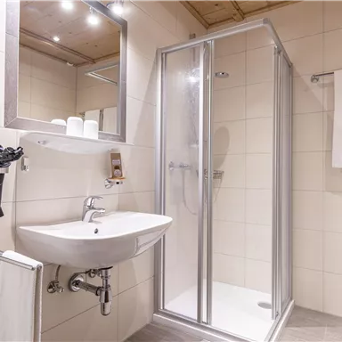 Ein modernes Badezimmer mit einer Dusche, einem Waschbecken und einem großen Spiegel. Handtücher und ein Föhn sind ebenfalls vorhanden.