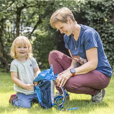 Eine Mutter sitzt mit ihrem kleinen Kind im Garten. Sie helfen sich gegenseitig, einen blauen Rucksack auszupacken.