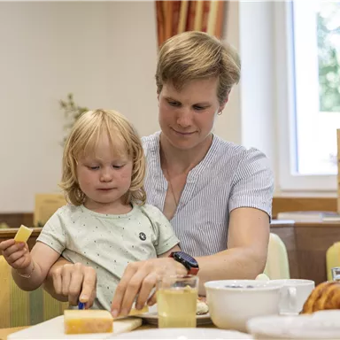 Eine Mutter und ihr kleines Kind sitzen an einem Tisch und genießen ihr Frühstück. Sie bereiten gemeinsam Essen vor, während im Hintergrund das gemütliche Ambiente einer Frühstücksgelegenheit zu sehen ist.