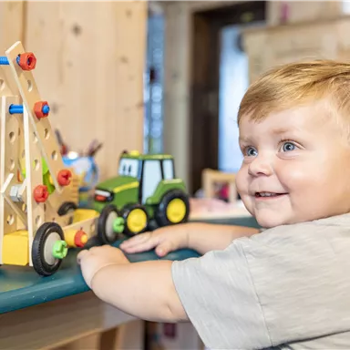Ein fröhlicher Junge spielt mit Holzspielzeug auf einem Tisch. Neben ihm stehen ein Spielzeugtraktor und ein Kran aus bunten Bausteinen.