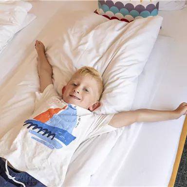Ein lächelnder Junge liegt entspannt auf einem Bett mit weißen Laken. Er trägt ein T-Shirt mit buntem Design und genießt die gemütliche Atmosphäre.