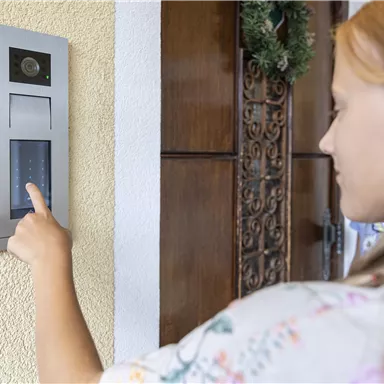 Eine Frau steht vor einer Haustür und drückt einen Türsummer. Im Hintergrund ist eine dekorative Tür zu sehen.