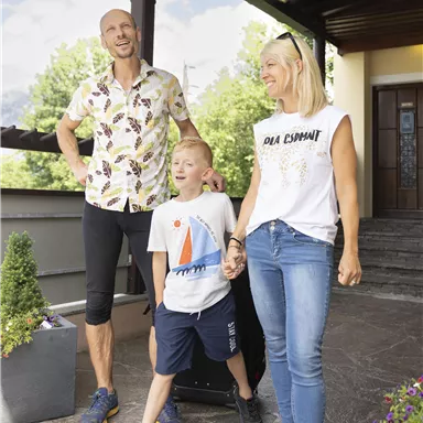 Eine Familie steht vor einem Haus mit einer freundlichen Atmosphäre. Der Vater trägt ein buntes Hemd, während die Mutter und der Sohn lässige Kleidung tragen und zusammen lachen.