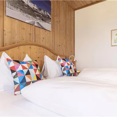 Ein gemütliches Schlafzimmer mit einem Holzpaneel und bunten Kissen. Auf der Wand hängt ein Kunstwerk, das eine Berglandschaft zeigt.