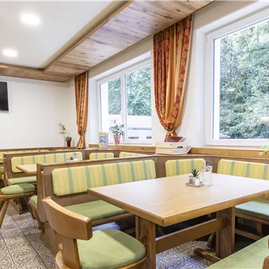 Ein gemütlicher Speisesaal mit Holzmöbeln und bunten Bänken. Große Fenster lassen viel Tageslicht herein und bieten einen Blick ins Grüne.