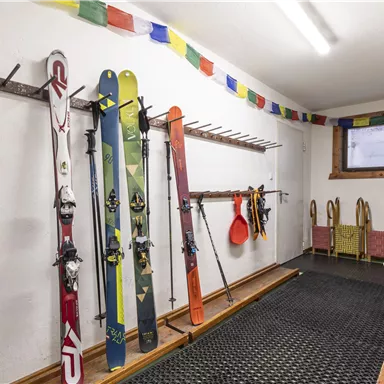 Ein Raum mit fünf Skiern an der Wand und einem Regal für Skischuhe. Bunte Wimpel schmücken die Decke.