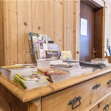Ein hölzerner Tisch mit stapelweise Broschüren und Prospekten. Im Hintergrund sieht man eine Holzvertäfelung und eine offene Tür.