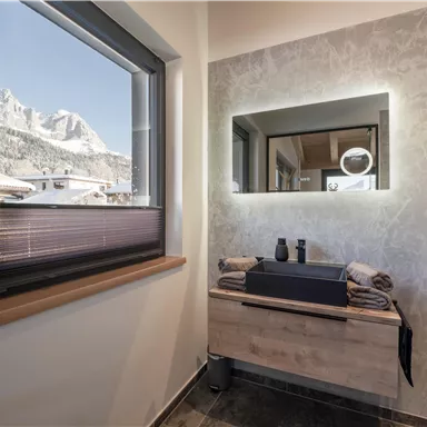 Ein modernes Badezimmer mit Fenster und Blick auf schneebedeckte Berge. Es gibt einen stilvollen Waschtisch mit Handtüchern und dekorativen Elementen.