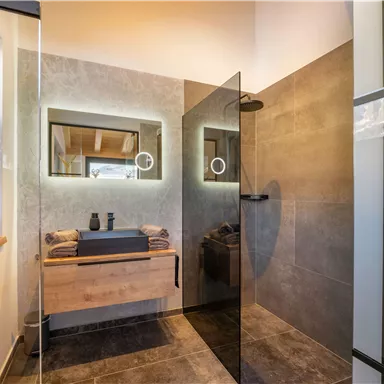 Ein modernes Badezimmer mit einer geräumigen Dusche aus Glas und stilvollen Spiegeln. Die Wände sind in grauen Tönen gehalten und es gibt einen Holzschrank mit Handtüchern.