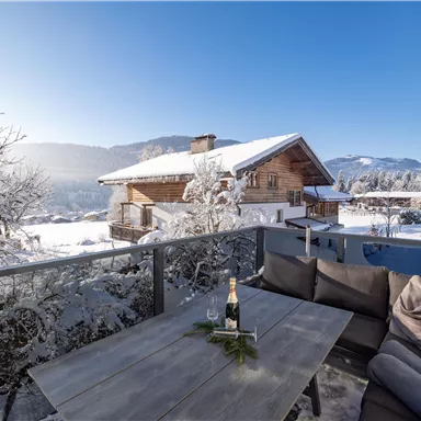 Eine gemütliche Terrasse mit einem Tisch und Sitzmöglichkeiten, umgeben von schneebedeckten Bergen und Häusern. Der klare, blaue Himmel verleiht der winterlichen Landschaft eine friedliche Atmosphäre.