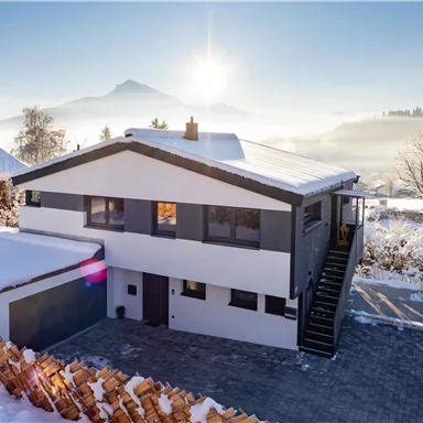 Ein modernes Haus im Schnee mit einem klaren blauen Himmel und der Sonne im Hintergrund. Die Landschaft ist ruhig und winterlich.