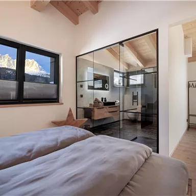 Ein modernes Schlafzimmer mit zwei Betten und großen Fenstern. Im Hintergrund sind schneebedeckte Berge und ein elegantes Badezimmer sichtbar.