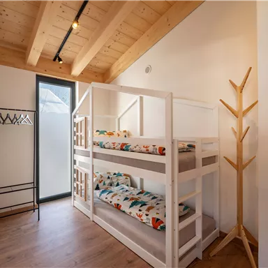 Ein helles Zimmer mit einem Etagenbett und bunten Bettwäsche. An der Wand steht ein Kleiderständer und ein großer Fenster bringt viel Licht hinein.