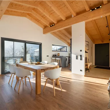 Ein modernes Wohnzimmer mit hohen Holzdecken und großen Fenstern. Ein Esstisch mit Stühlen und eine dekorative Rehfigur sorgen für eine gemütliche Atmosphäre.