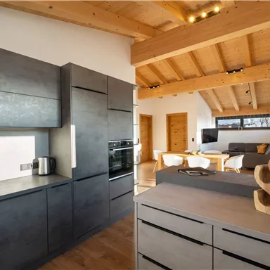 Eine moderne Küche mit dunklen Schränken und Holzdecke. Im Hintergrund ist ein gemütlicher Wohnbereich sichtbar.