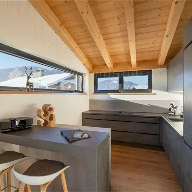 Eine moderne Küche mit Holzdecke und großen Fenstern, die viel Licht hereinlassen. Die Möbel sind in neutralen Farben und bieten eine elegante, schlichte Ästhetik.