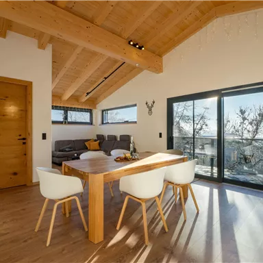 Ein modernes Wohnzimmer mit Holzdecke und großen Fenstern. Ein Esstisch mit weißen Stühlen steht vor einer Glasschiebetür, die einen Blick nach draußen bietet.