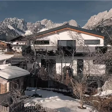 Ein modernes Haus in einer schneebedeckten Landschaft mit Bergen im Hintergrund. In der Nähe steht eine traditionelle Holzhütte, umgeben von Bäumen.