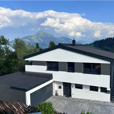 Ein modernes Haus mit flachem Dach und großen Fenstern. Im Hintergrund sind Berge und ein blauer Himmel zu sehen.