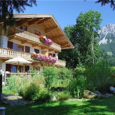 Ein schönes Holzhaus mit Balkonen, geschmückt mit Blumen. Im Hintergrund sind mit Bäumen bewachsene Berge zu sehen.