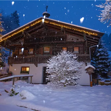 Ein gemütliches Holzhaus im Schnee, umgeben von verschneiten Tannen. Sanfte Schneeflocken fallen bei Nacht, erleuchtet von warmen Lichtern.