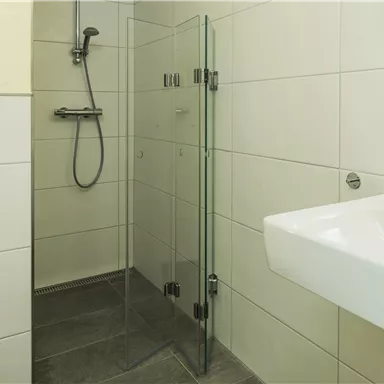 Ein modernes Badezimmer mit einer Glasdusche und einem Waschbecken. Die Wände sind mit hellen Fliesen gefliest.