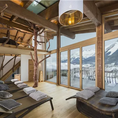 Ein modernes, helles Wohnzimmer mit Holzdekor, großen Fenstern und Blick auf die Berge. Es gibt Liegen und eine gemütliche Atmosphäre.