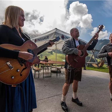 Eine Gruppe von Musikern spielt draußen. Sie tragen traditionelle Kleidung und die Atmosphäre ist festlich.