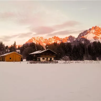 Eine malerische Winterlandschaft mit schneebedeckten Feldern und Holzhäusern. Im Hintergrund erstrahlen die Berge im Sonnenlicht.