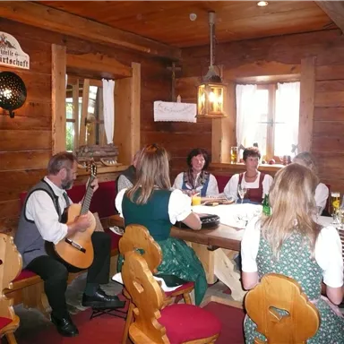 Eine gemütliche Stube mit Holzdekor und traditionellen bayerischen Trachten. Menschen sitzen am Tisch und genießen Musik und Geselligkeit.