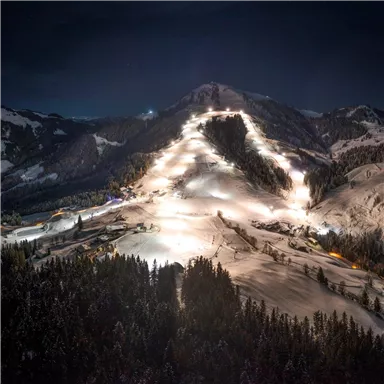 Eine winterliche Berglandschaft bei Nacht mit beleuchteten Skipisten. Im Vordergrund sind verschneite Bäume zu sehen.