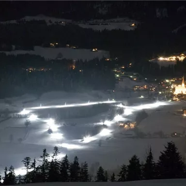 A illuminated winter landscape at night, with snow-covered hills and a small village in the background. The lights brighten the surroundings and create a cozy atmosphere.