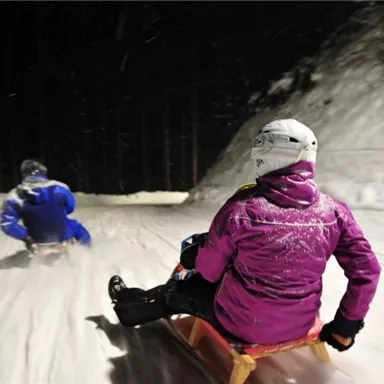Two sledders are going down a snow-covered slope at night. The surroundings are dark and the snow is flying up as they speed along.