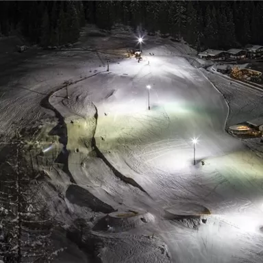 A snow-covered ski slope at night, illuminated by floodlights. In the background, cozy cabins can be seen, surrounded by pine trees.