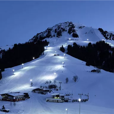 Eine verschneite Berglandschaft im Abendlicht. Lichter erhellen die Piste und das Tal darunter.