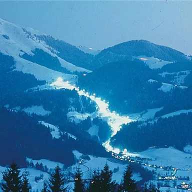 A wintry mountain landscape with snow-covered hills. Gentle lights illuminate the slopes and the village in the valley.